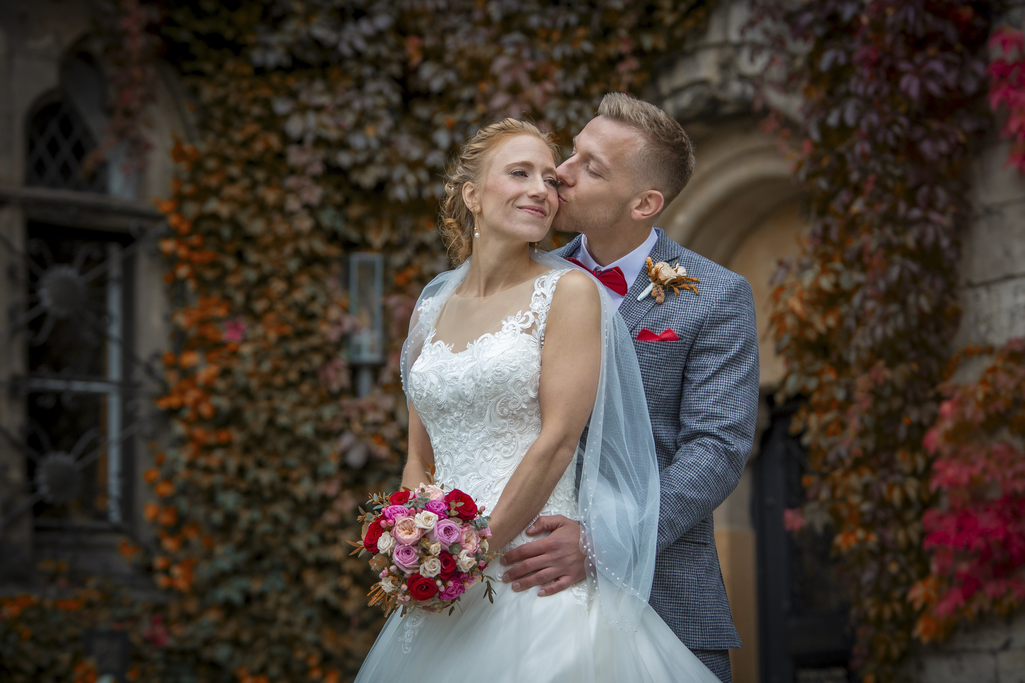 Hochzeitsfotograf - Auf diesem Foto, das ich an einem herrlichen Herbsttag aufgenommen habe, strahlen die Braut und der Bräutigam vor einem Hintergrund, der in den warmen Farben der Saison leuchtet. Die Braut, in ihrem strahlenden Kleid, lächelt glückselig, während der Bräutigam liebevoll seine Lippen an ihre Wange schmiegt. Sein schickes, graues Karomuster-Anzug und die rote Fliege kontrastieren charmant mit dem lebendigen Blumenstrauß, den die Braut hält. Die altehrwürdige Architektur und das reich gefärbte Laub verleihen dem Bild eine romantische und zeitlose Qualität. Es war ein besonderer Augenblick, den ich festhalten durfte – ein Bild voller Liebe und Verbundenheit. - Mariana Siegert