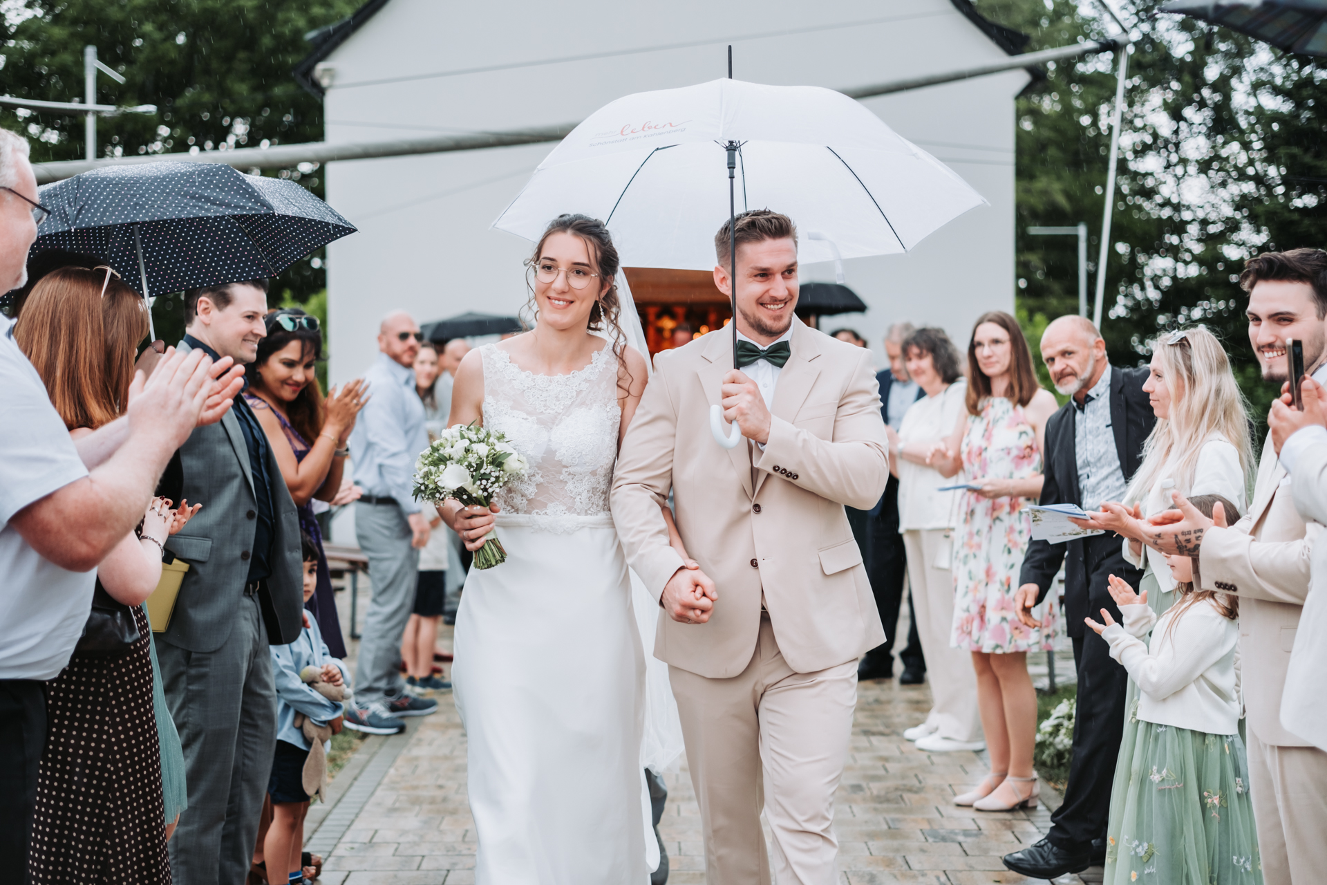 Hochzeitsfotograf: Brautpaar geht bei Regen unter dem Schirm durch das Spalier der applaudierenden Gäste am Kahlenberg in Wien - Pengoo Weddings