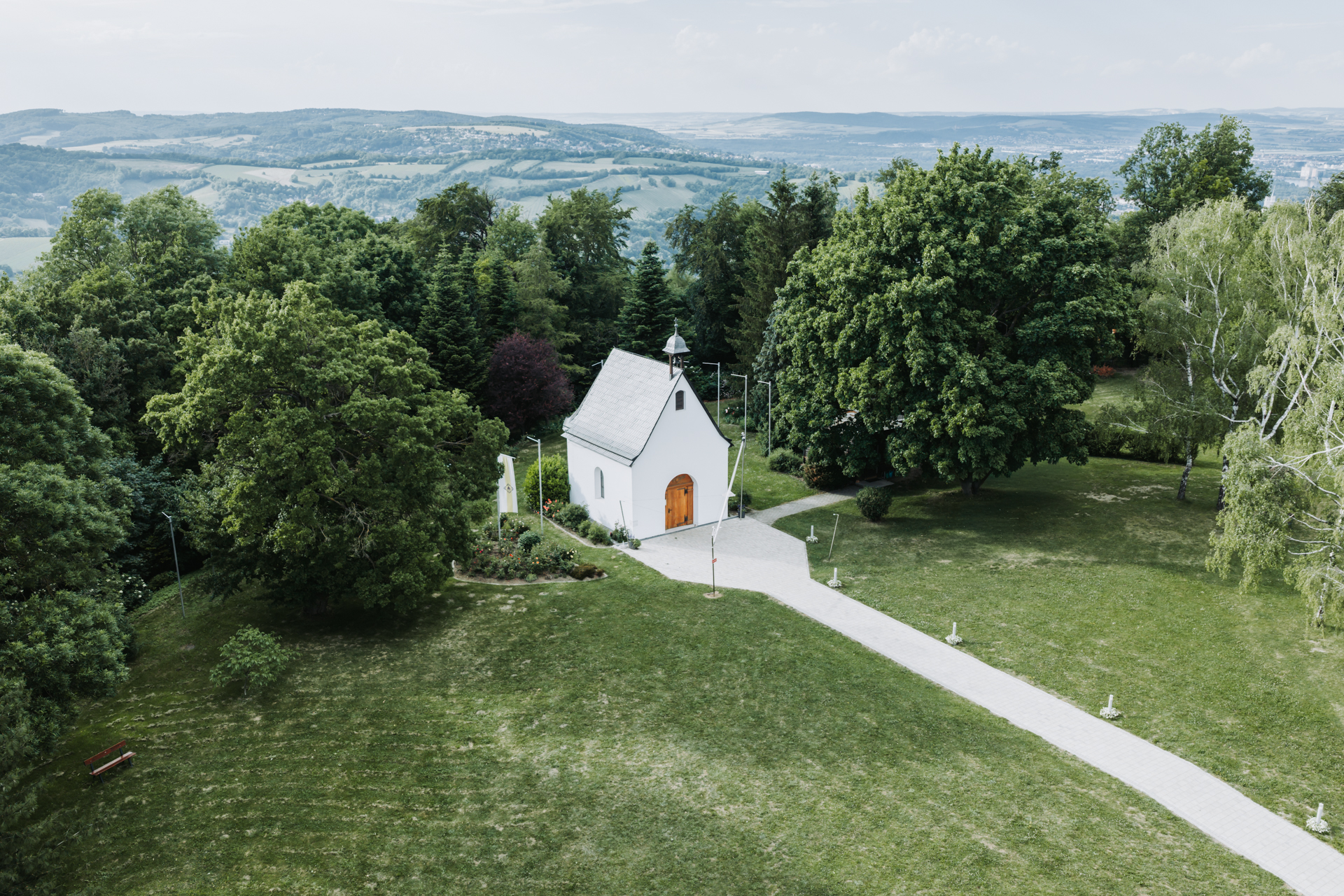 Hochzeitsfotograf: Luftaufnahme einer kleinen weißen Kapelle zwischen grünen Bäumen mit Blick über Wien am Kahlenberg - Pengoo Weddings