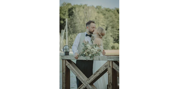 Hochzeitsfotos - Naumburg (Burgenlandkreis) - In dem Bild halte ich einen wunderschönen Moment von Anne und Max an ihrem Hochzeitstag fest. Sie stehen eng umschlungen auf einer alten Holzbrücke, umgeben von der ruhigen Natur. Max, in einem stilvollen Anzug mit weißer Weste und dunkler Fliege, gibt Anne, die in einem eleganten, ärmellosen Hochzeitskleid strahlt, einen liebevollen Kuss. Sie hält einen prachtvollen Strauß aus grünen und weißen Blumen. Die sanfte Beleuchtung umspielt das Paar und betont die Zärtlichkeit dieses intimen Moments. Es war eine Freude, die Gefühle und die Verbundenheit, die sie an diesem besonderen Tag teilten, einzufangen und für die Ewigkeit zu bewahren. - Mariana Siegert